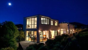 A modern two-story house with large windows and a deck, surrounded by greenery.
