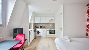 Une chambre d’hôtel compacte comprenant un lit, une petite table, une chaise rouge, un téléviseur, un lave-linge et une kitchenette.