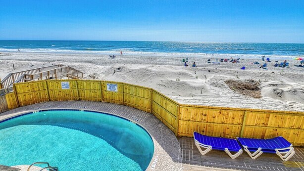 A beachside pool area with a wooden deck, a pool, and lounge chairs.
