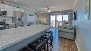 A modern kitchen with a large island, bar stools, and a view of the ocean through the sliding glass door.