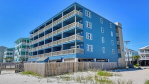 A multi-story beachfront hotel with balconies, a sandy beach in front, and a wooden fence.