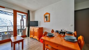 Een eethoek met een houten tafel en stoelen, een televisie en zicht op een besneeerd landschap door een groot raam.