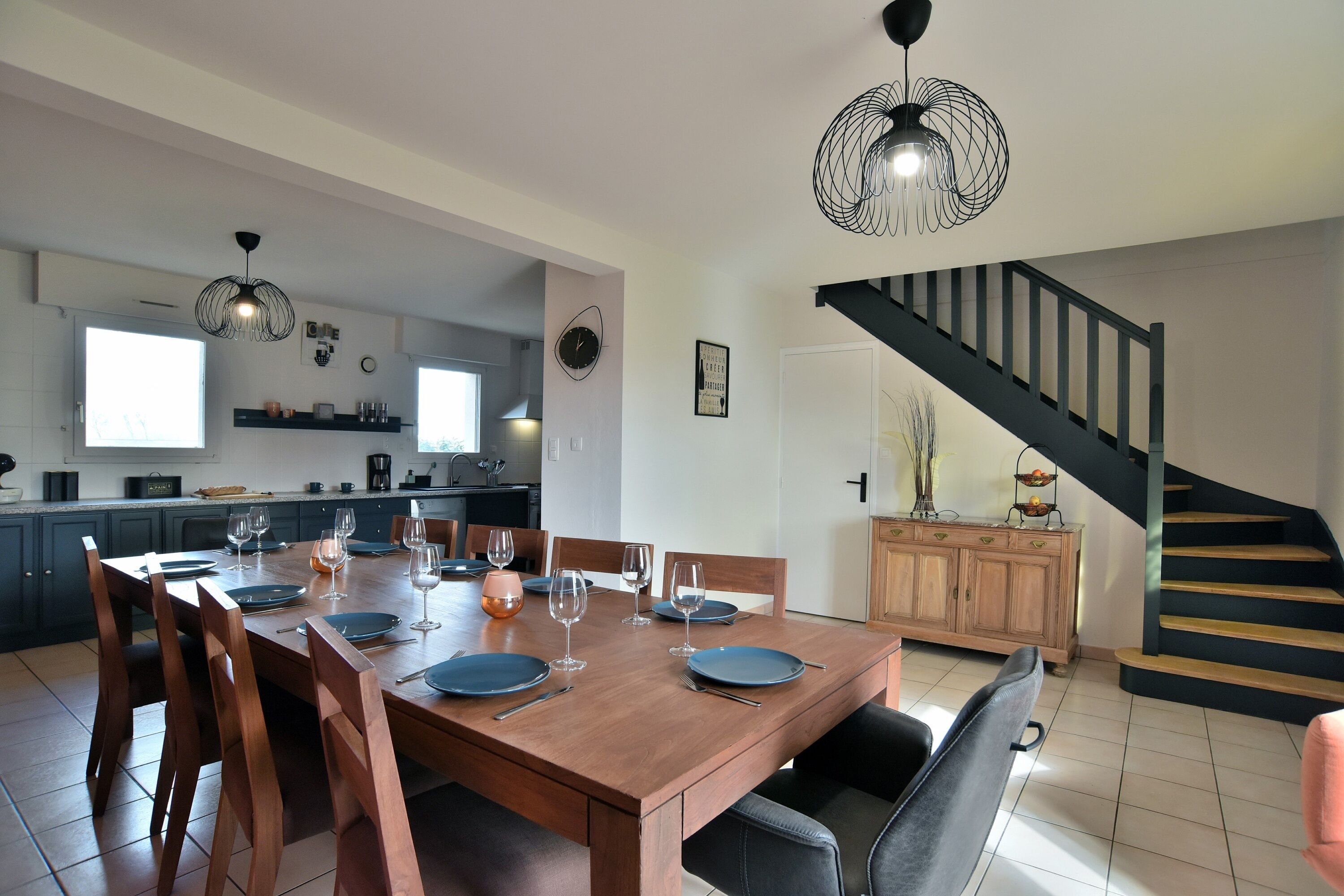 Een moderne keuken met donkere kastjes, een houten eettafel gedekt voor vier personen, en een trap met een zwarre leuning.