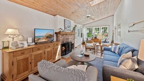 A spacious living room with a fireplace, wooden ceiling, and a large window offering a view of the outdoors.