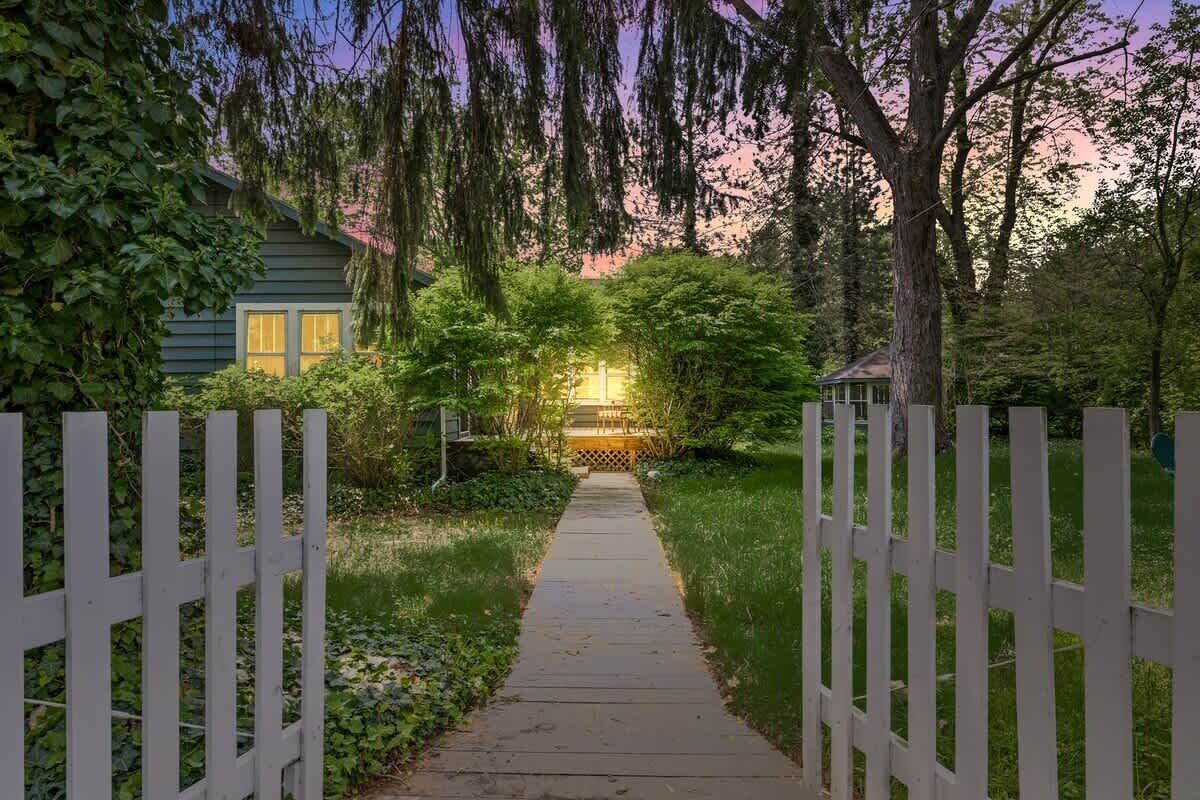A wooden pathway leading to a house with a white picket fence, surrounded by greenery and trees.