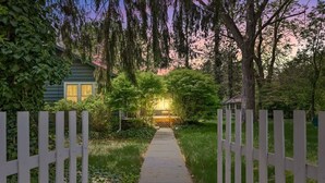 A wooden pathway leading to a house with a white picket fence, surrounded by greenery and trees.