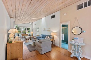 A spacious living room with a wooden ceiling, a blue sofa, a striped armchair, and a round table with a lamp.