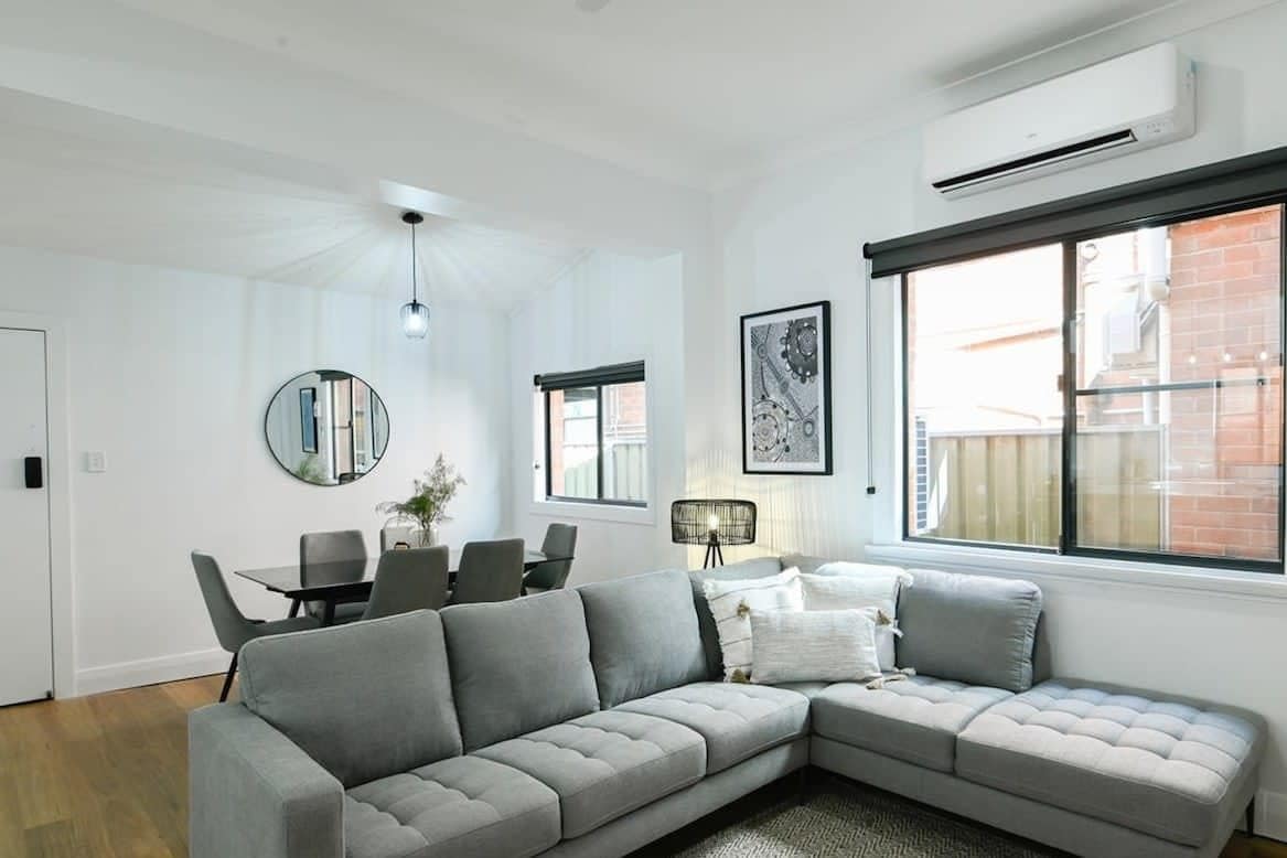 A modern living room with a grey sectional sofa, a dining table with chairs, a round mirror, and artwork on the walls.