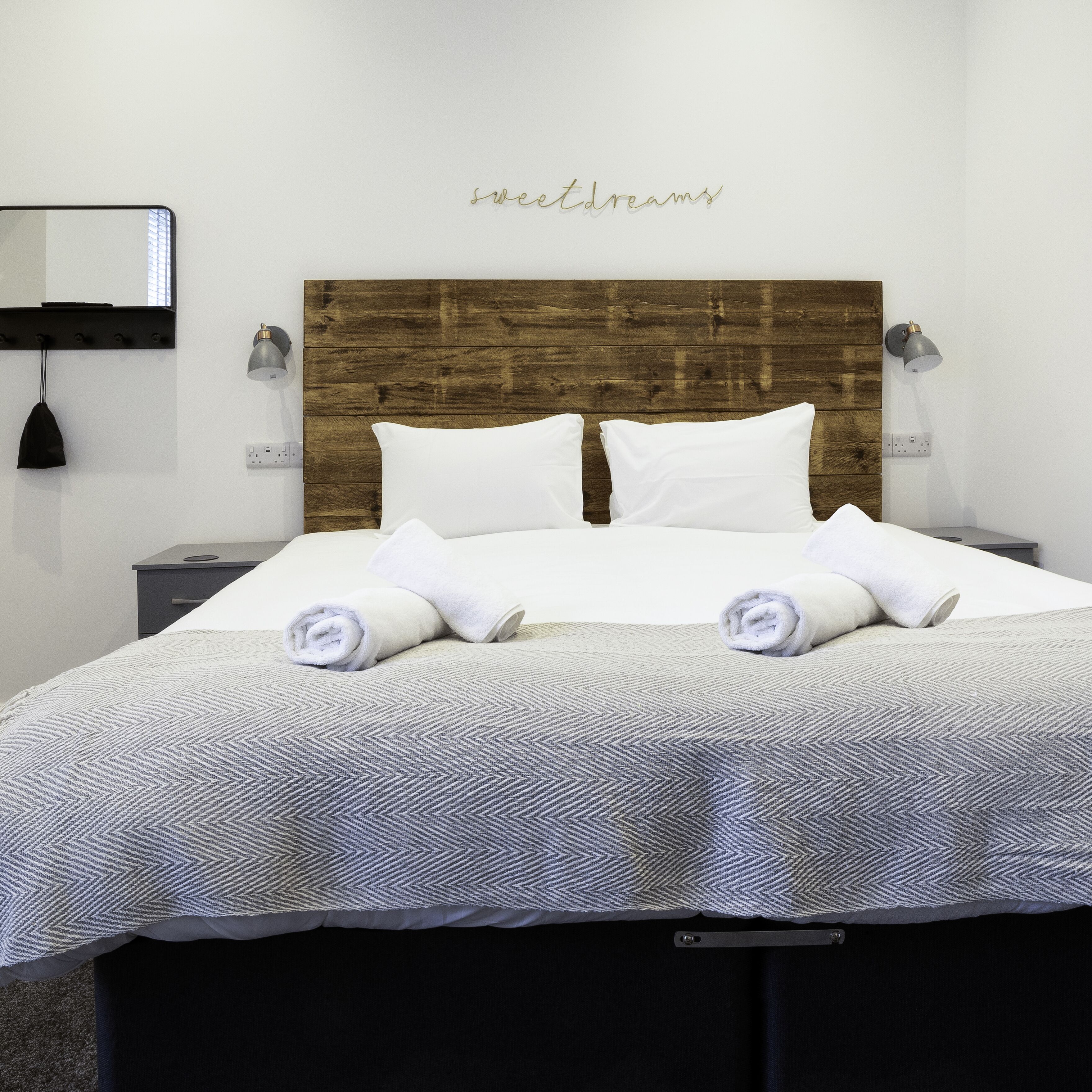A neatly made bed with white linens and two rolled towels, a wooden headboard, bedside lamps, and a wall-mounted light fixture.