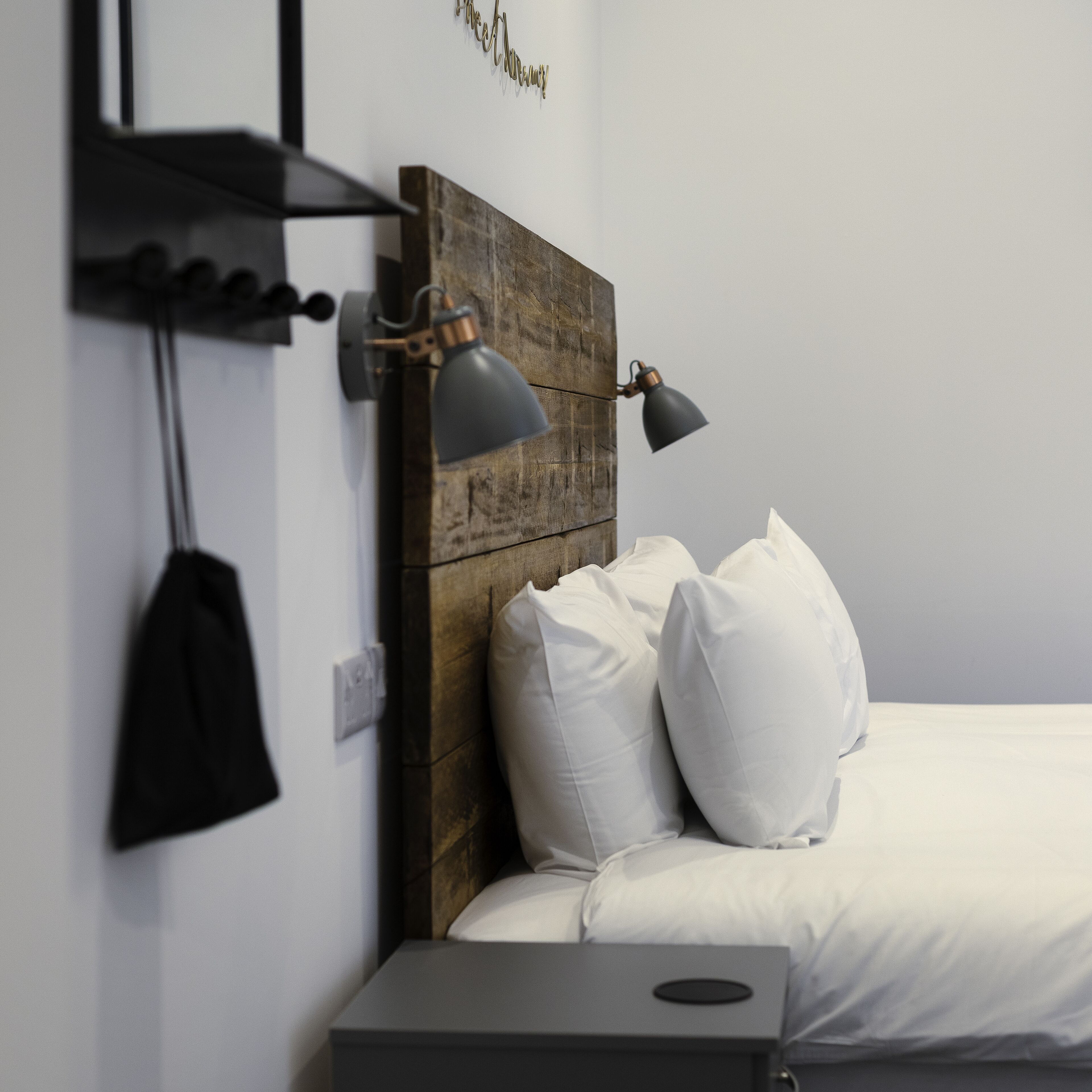 Una cama bien hecha con almohadas blancas y un cabecero de madera. Hay una mesita de noche junto a la cama y una lámpara fijada a la pared.