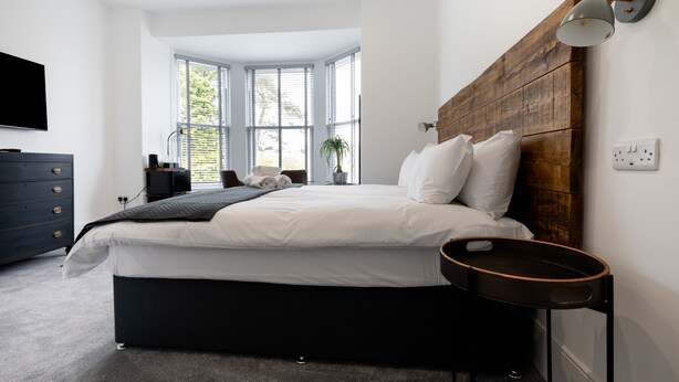 A modern bedroom with a large bed, a wooden headboard, a bedside table, a TV, and a window with blinds. - Park View (Falmouth)