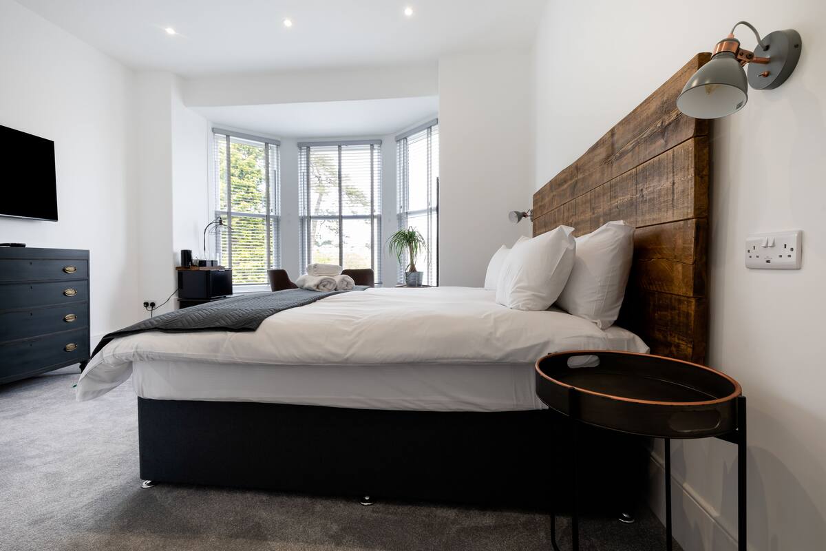 A modern bedroom with a large bed, a wooden headboard, a bedside table, a TV, and a window with blinds.