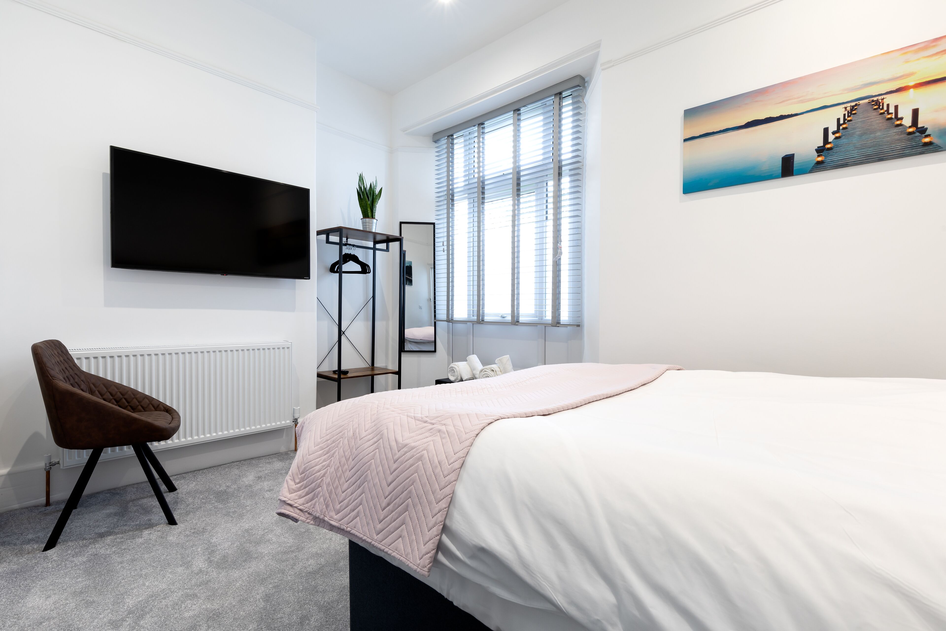 Quarto com cama, cadeira, televisão na parede, janela com persianas e um quadro na parede.