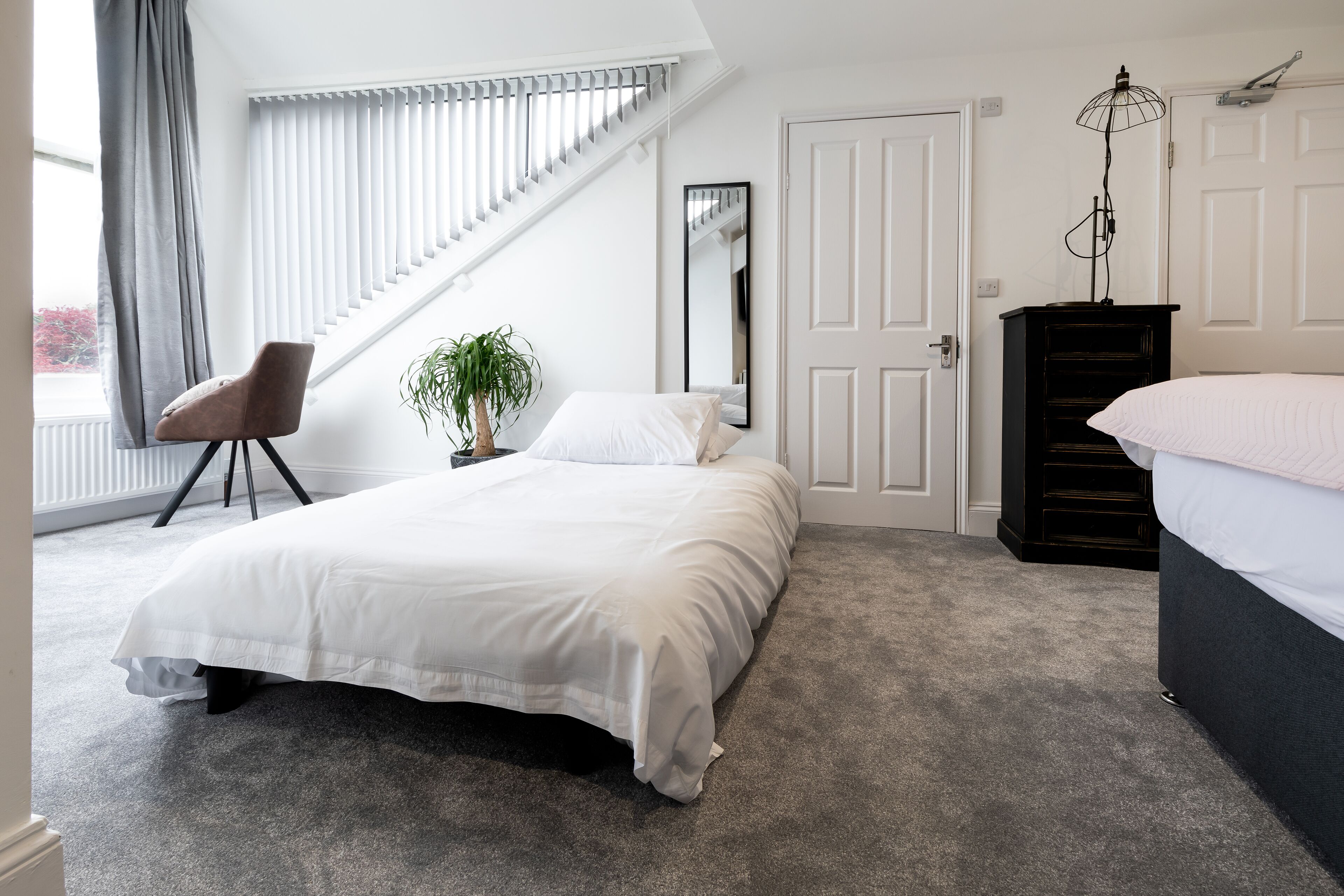 A bedroom with a large bed, a chair, a dresser, a plant, and a window with blinds.