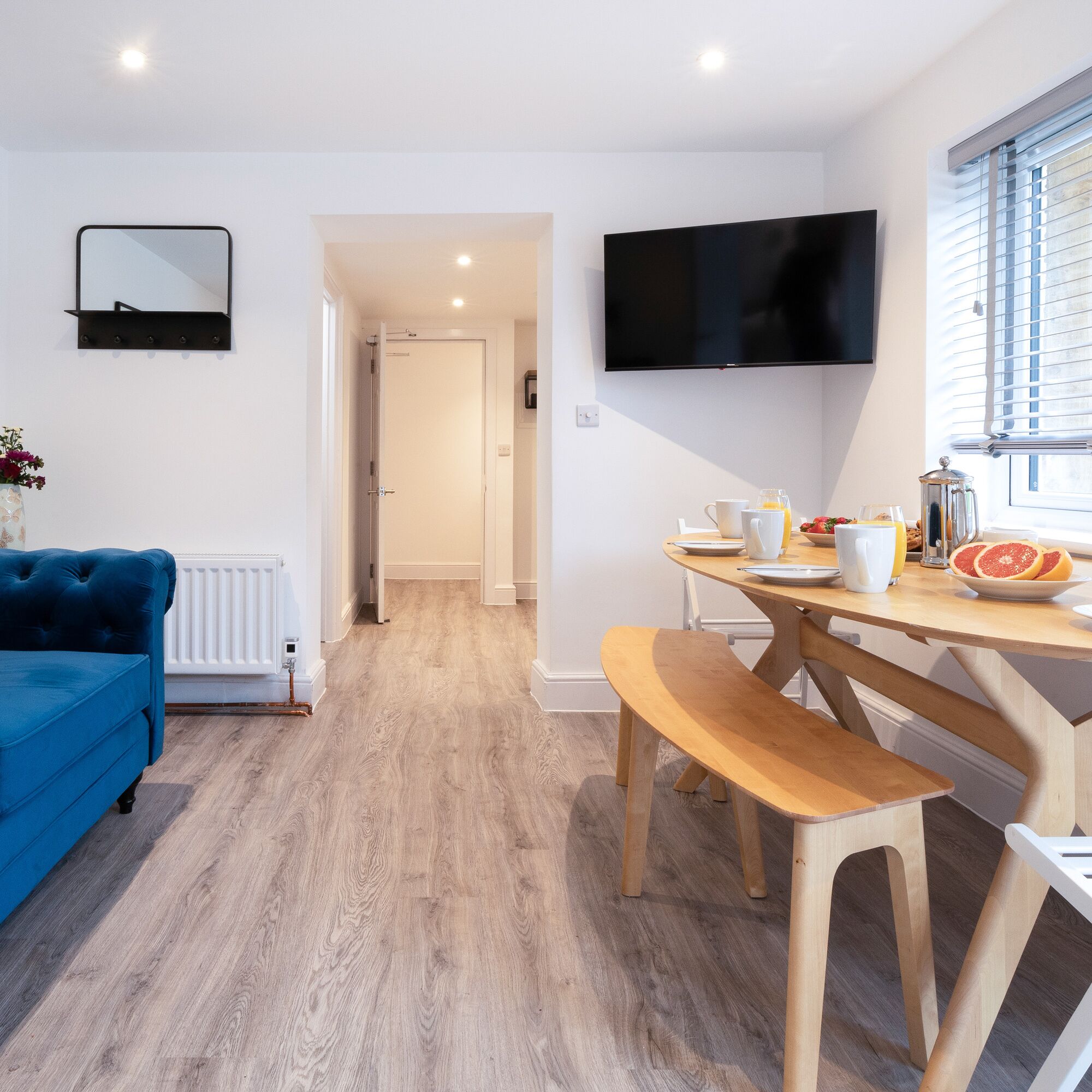 A modern living room with a blue sofa, a wooden dining table with benches, a flat-screen TV mounted on the wall, and a window with blinds.