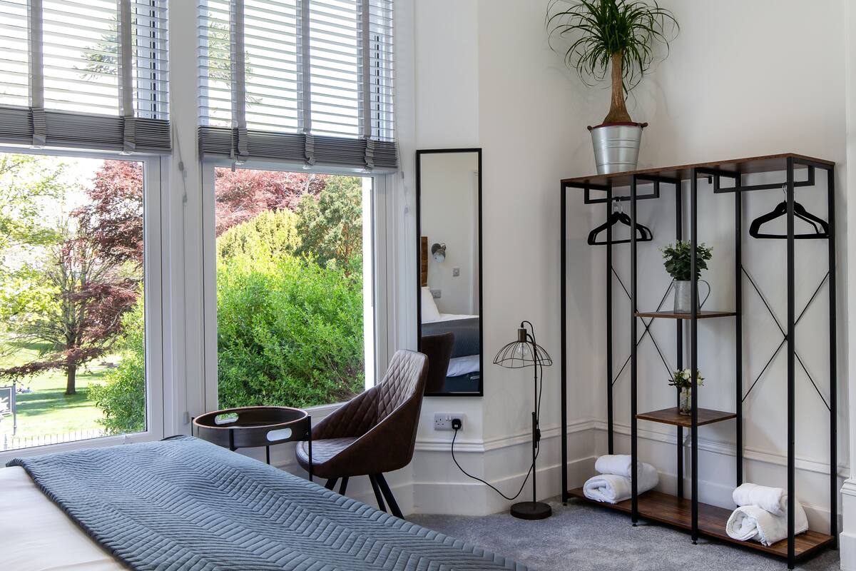 A bedroom with a large window, a bed, a chair, a mirror, a plant, and a metal shelf with hangers.
