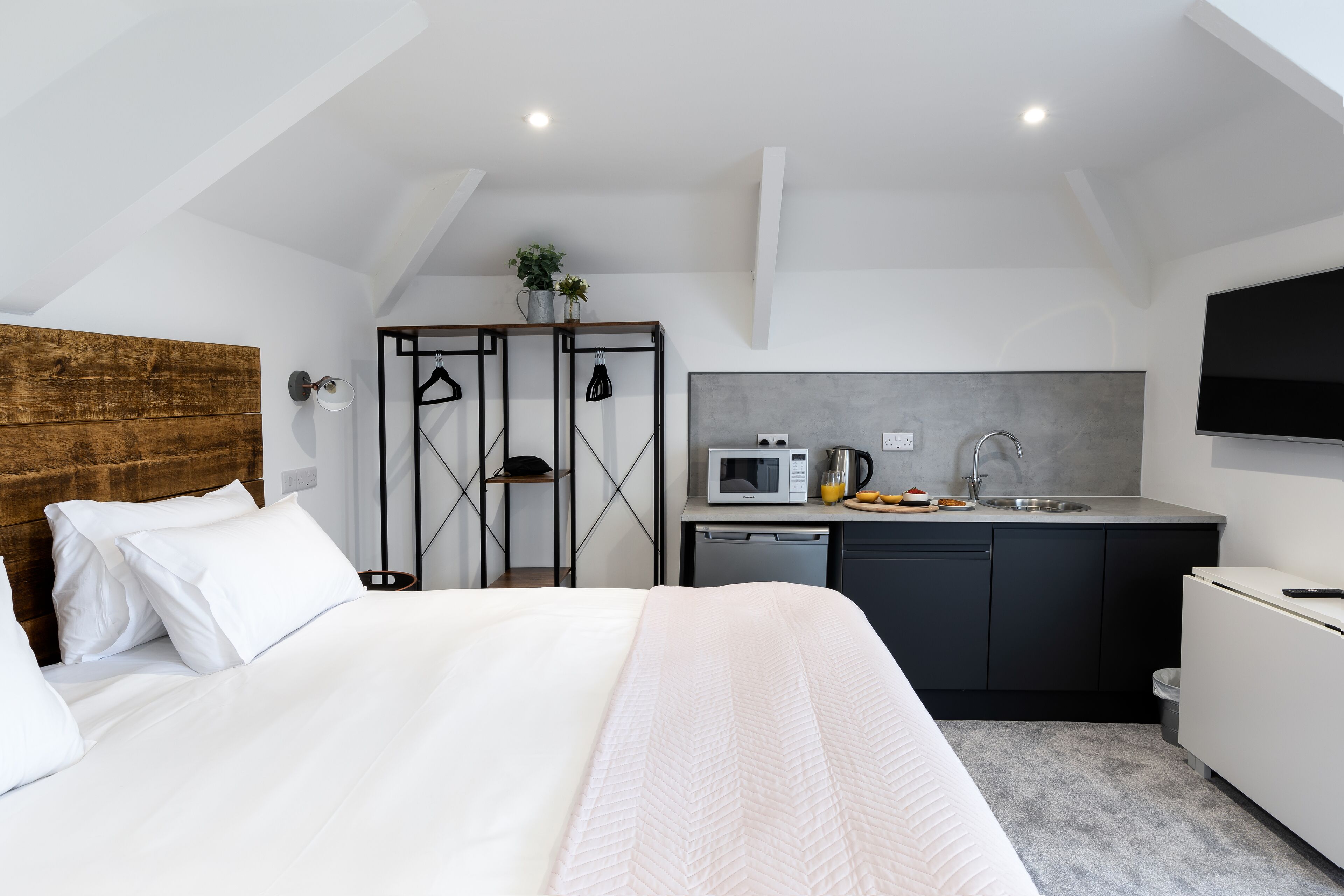 A modern hotel room with a large bed, a kitchenette, and a TV mounted on the wall.