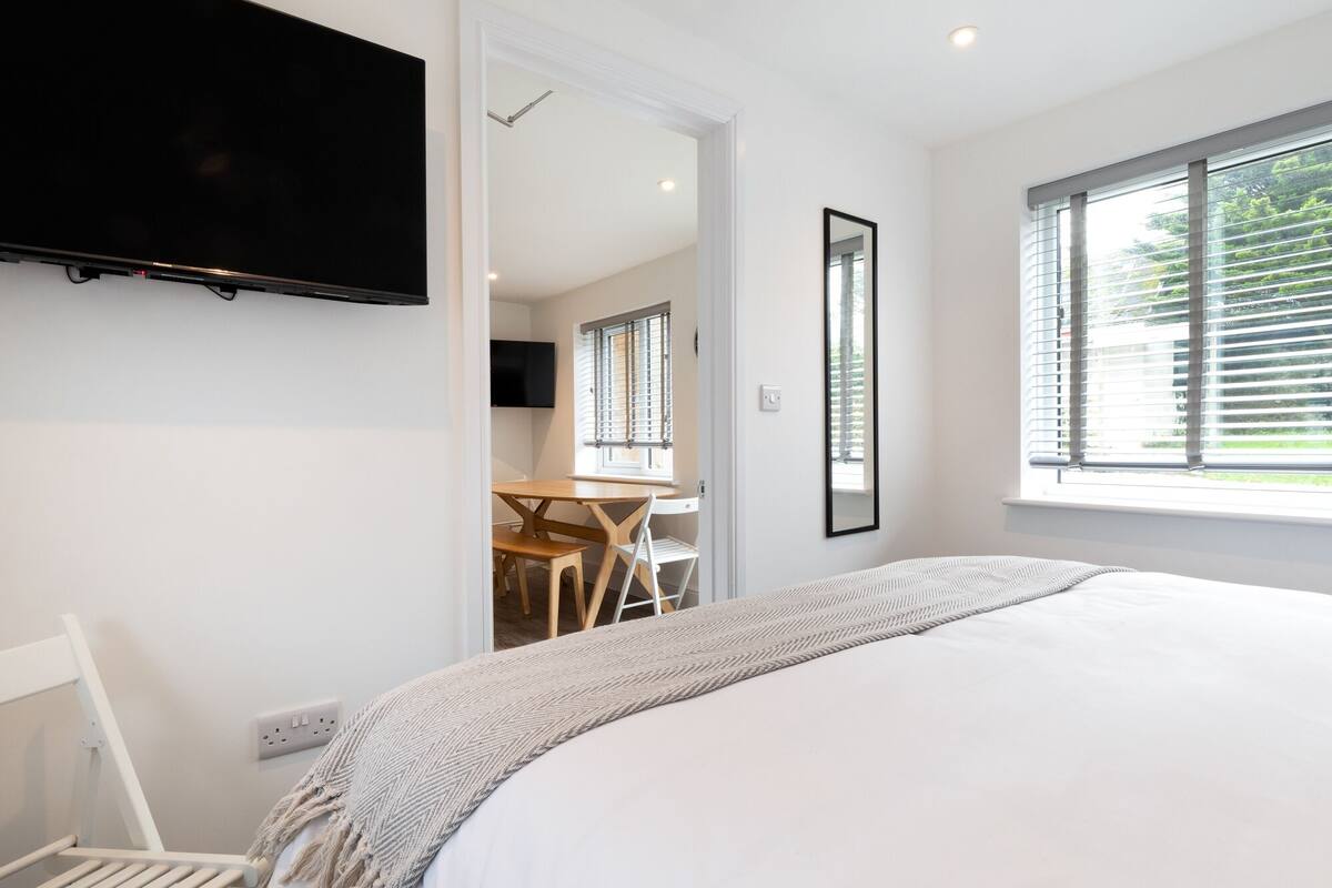 A bedroom with a bed, a TV mounted on the wall, a window with blinds, and a door leading to a dining area.