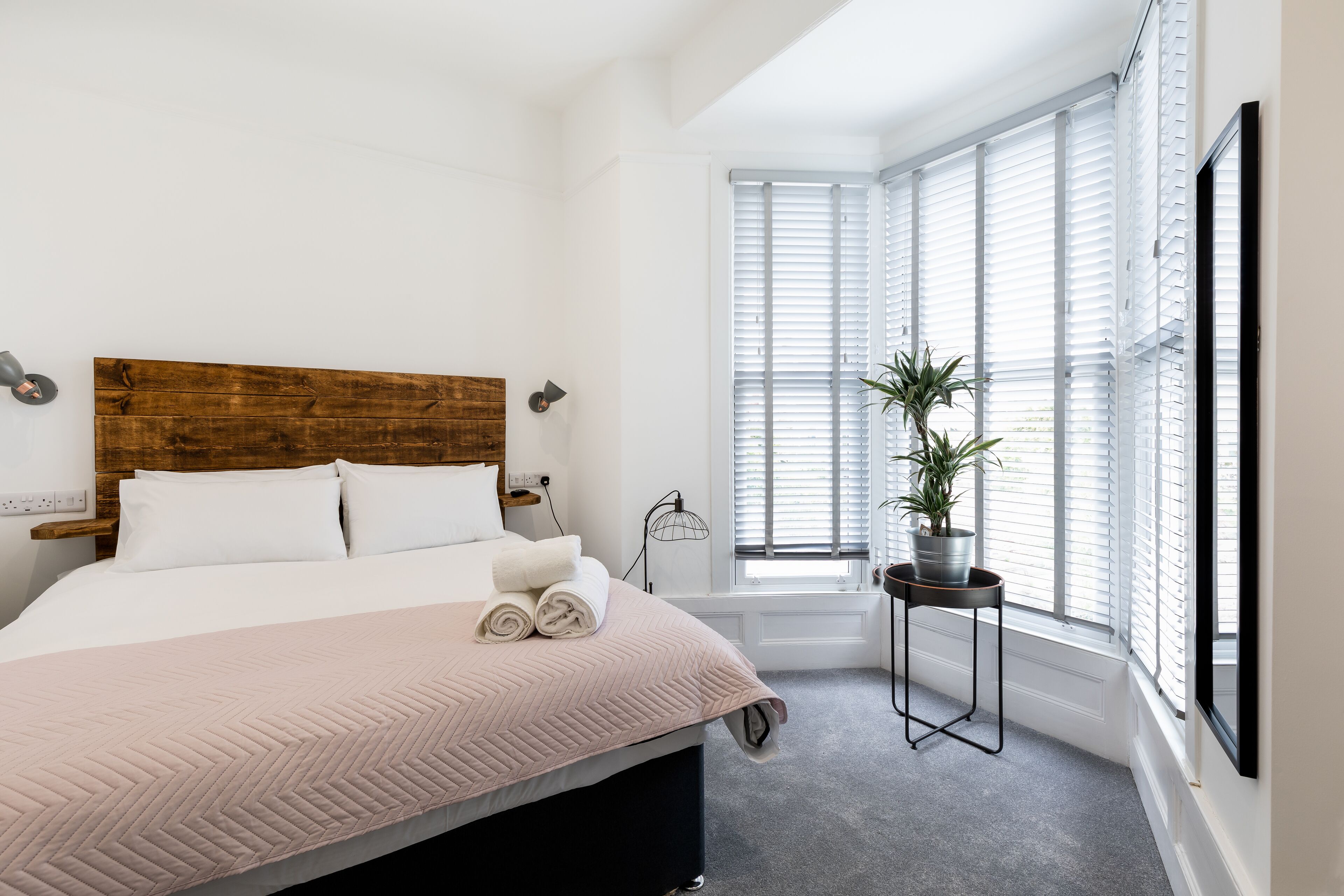 A bedroom with a wooden headboard, a bed with rolled towels, a potted plant, and a window with blinds.