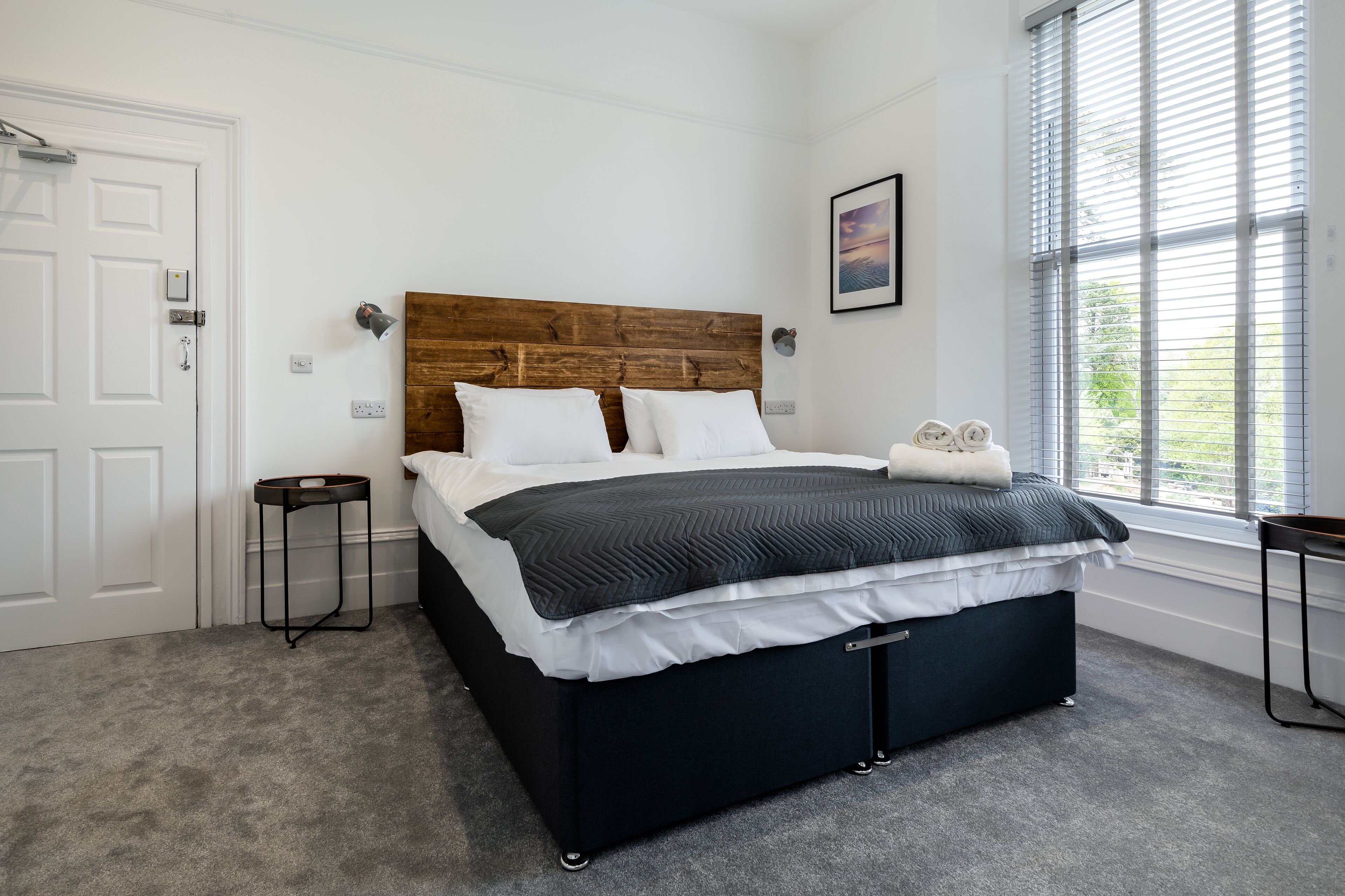 Un dormitorio con cama, mesitas de noche, una ventana con persianas y una puerta.