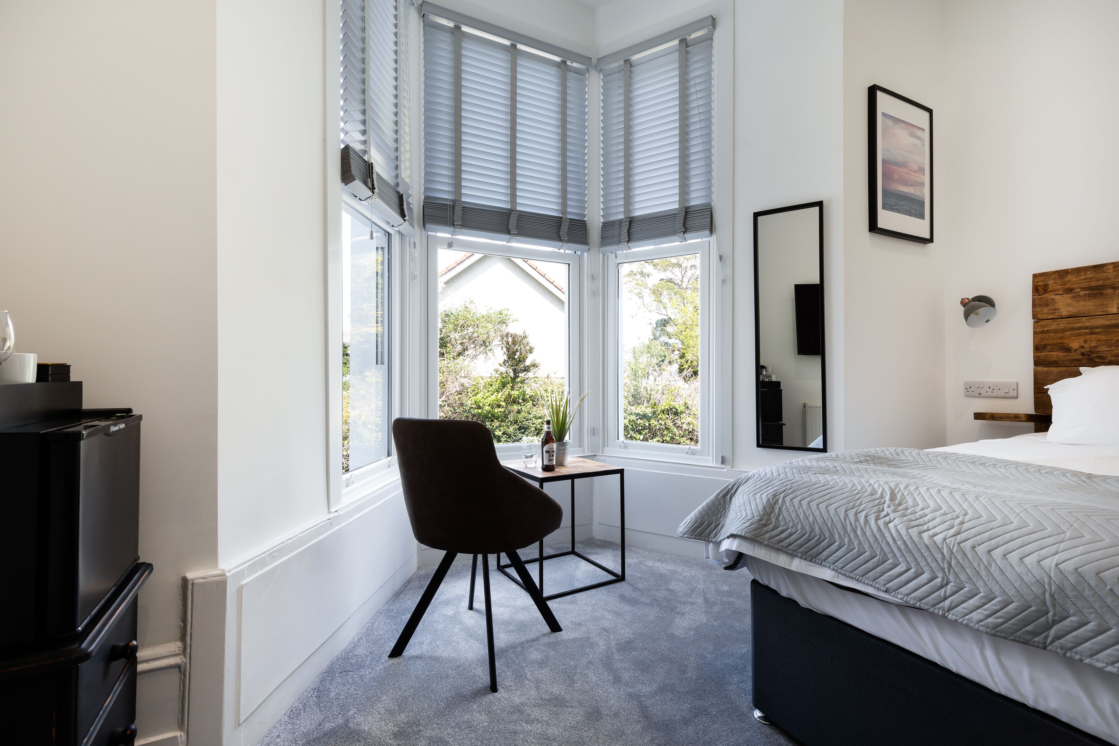 Une chambre à coucher comprenant un lit, un bureau, une chaise et une grande fenêtre avec des stores.