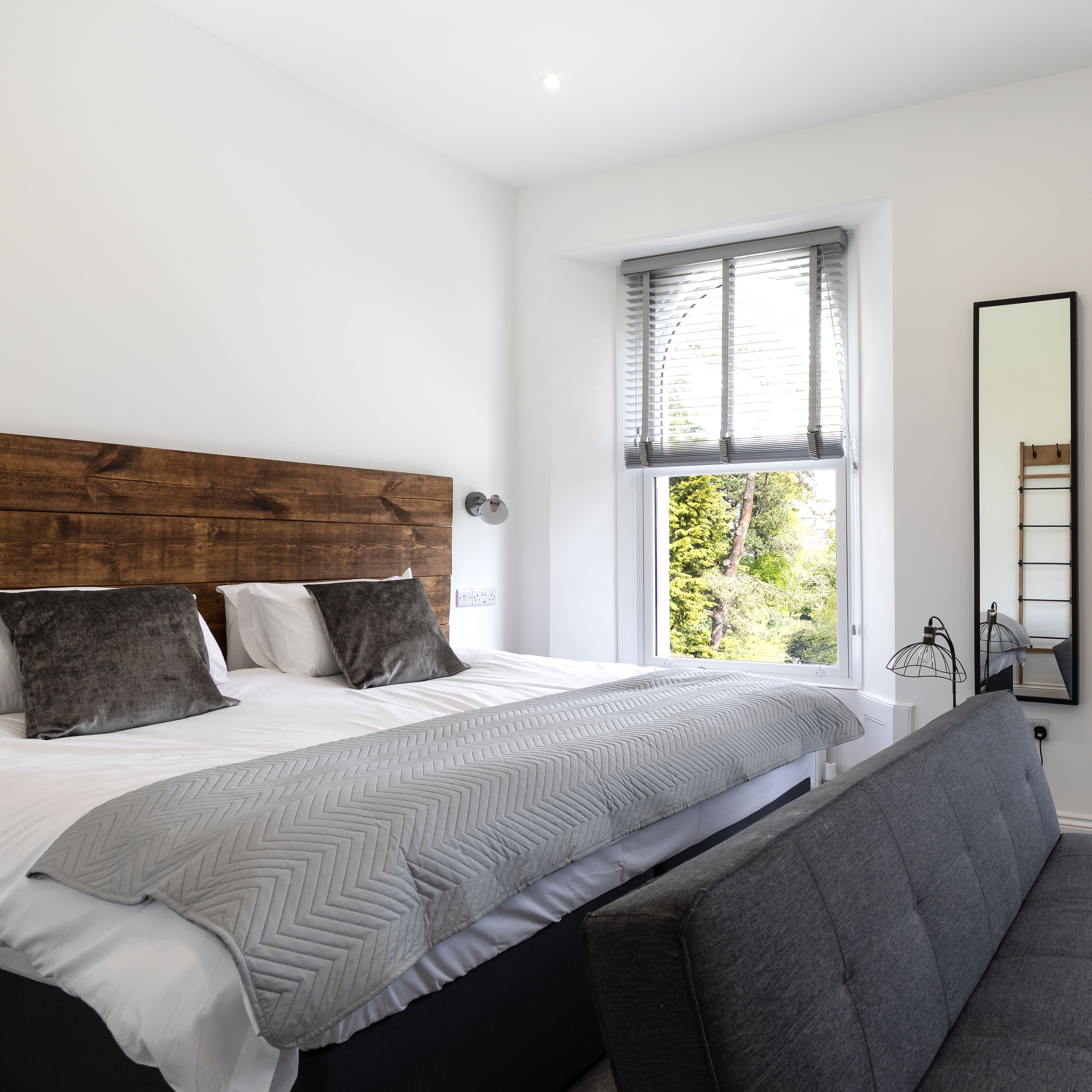 Een moderne slaapkamer met een houten hoofdbord, een groot bed met grijze beddengoed, een grijze bank, een raam met jaloezie ën en een spiegel aan de muur.