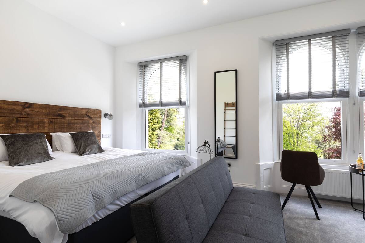 A bedroom with a large bed, a gray sofa, and two windows with blinds.