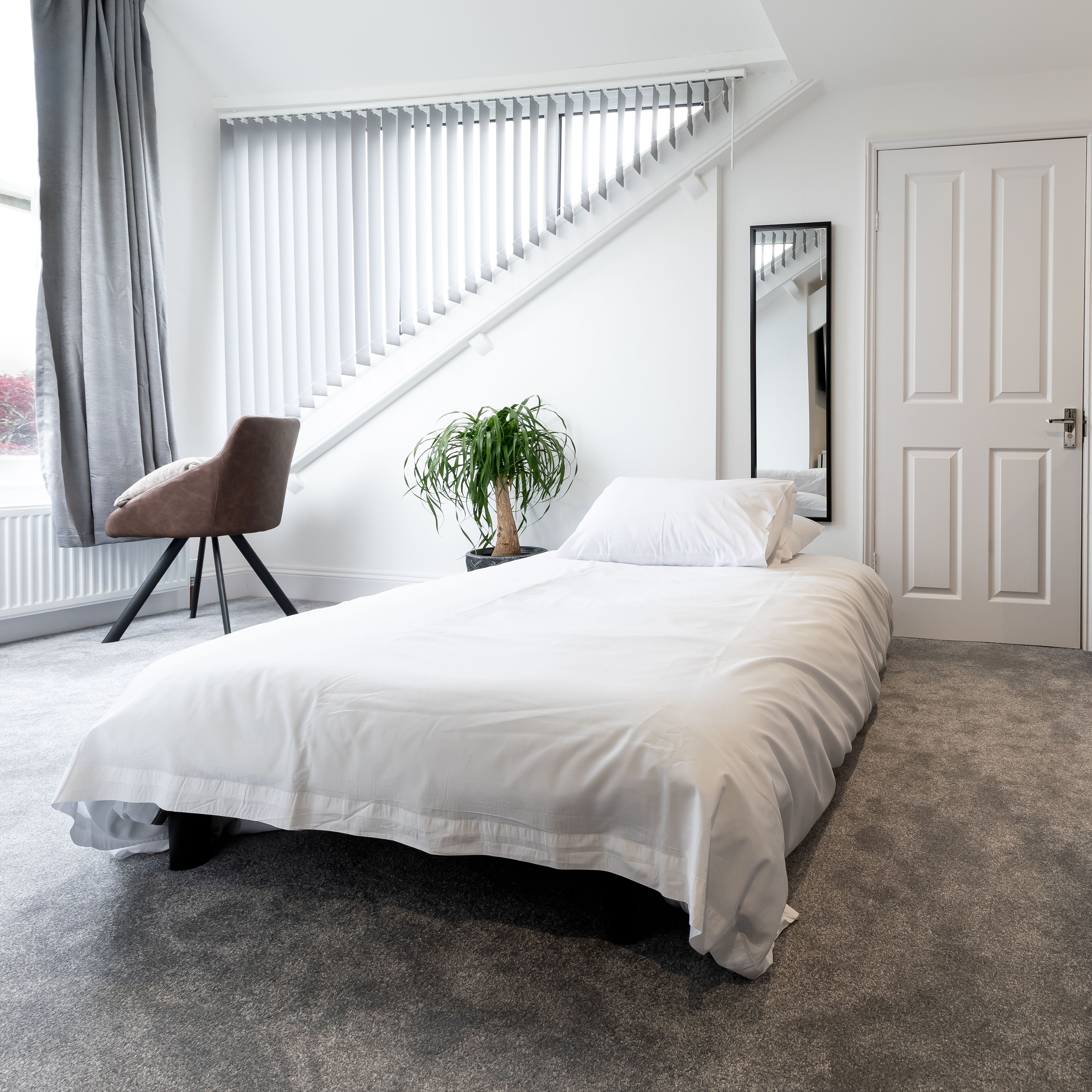 Een slaapkamer met een bed, een stoel, een plant, een raam met jaloezieën en een deur.