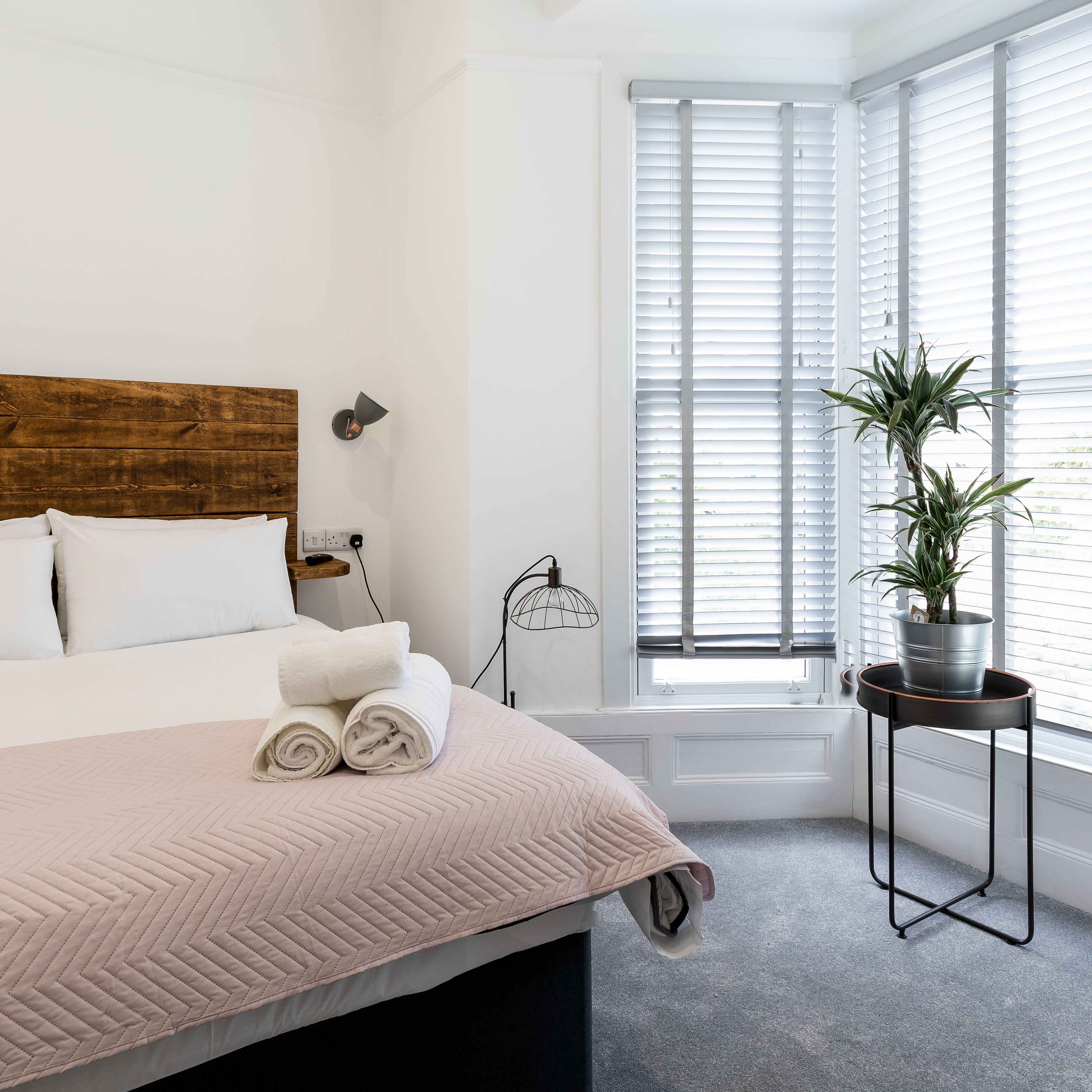 A neatly made bed with rolled towels, a wooden headboard, a bedside lamp, and a potted plant by a window with blinds.