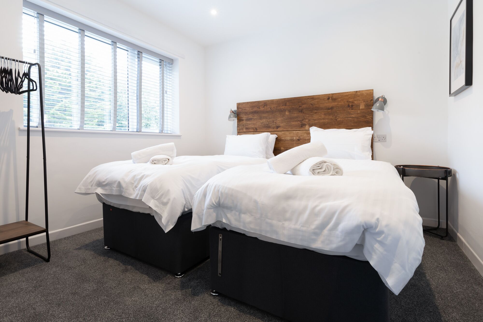 Une chambre double avec deux lits, une tête de lit en bois, une petite table de chevet et une fenêtre avec des stores.