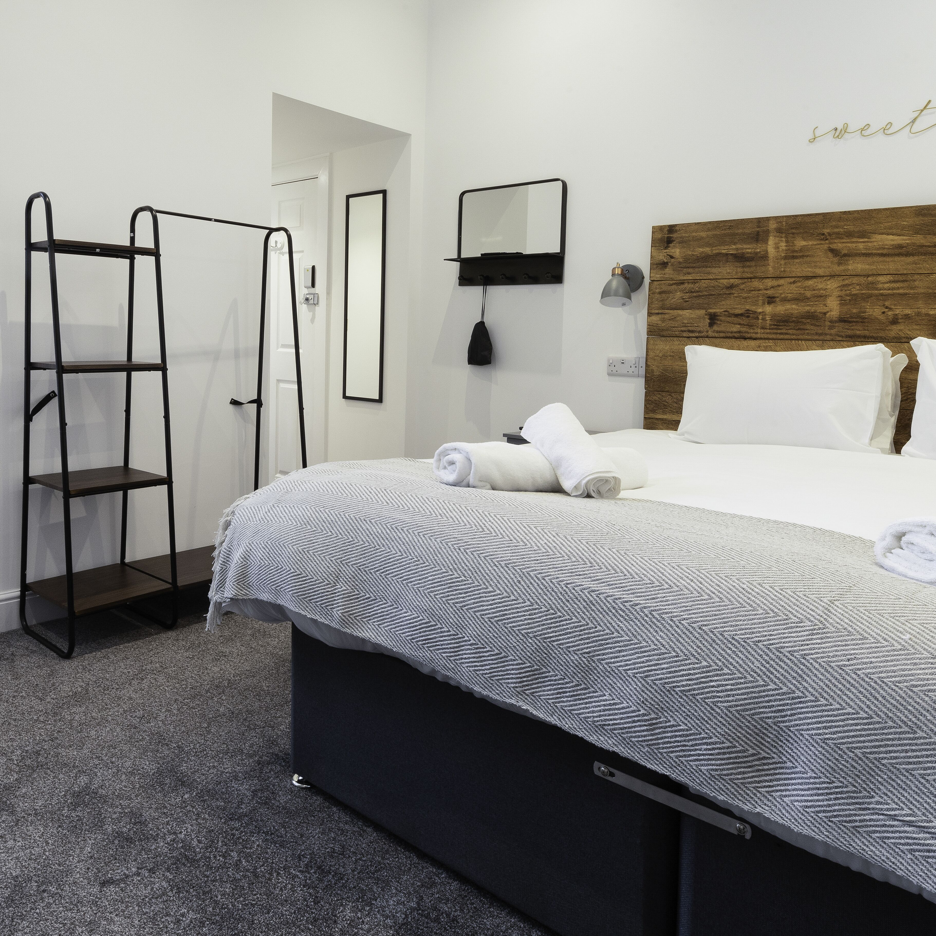 A hotel room with a bed, a wooden headboard, a ladder, a wall-mounted mirror, and a bedside lamp.