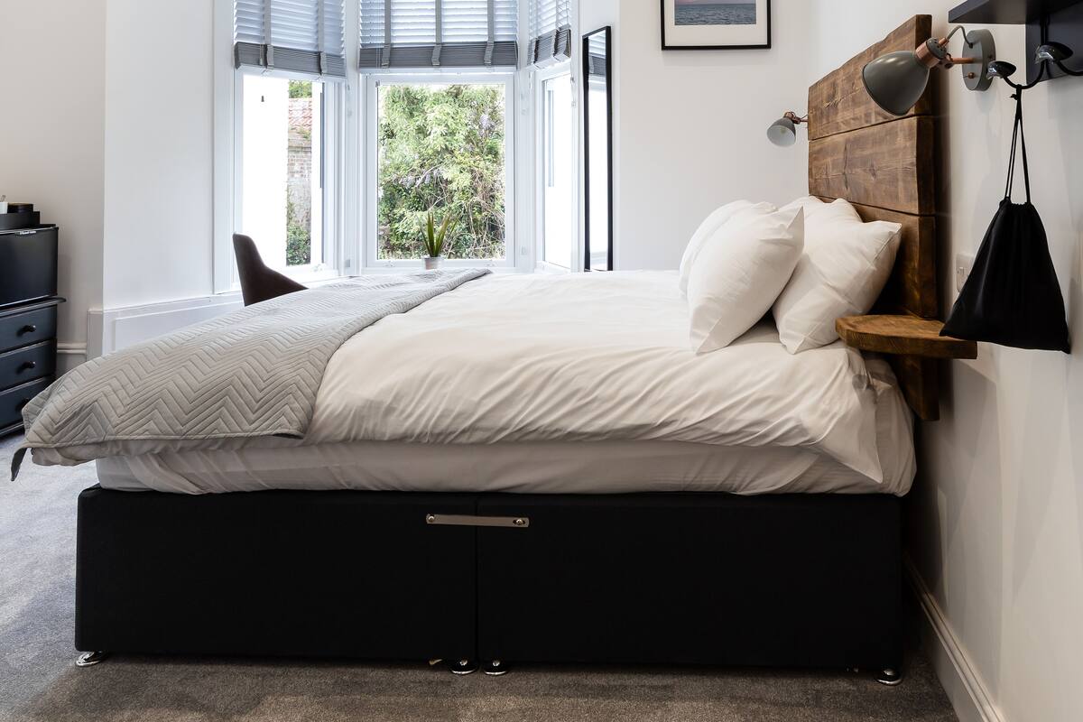 A bedroom with a large bed, a wooden headboard, a nightstand, a desk, and a window with blinds.