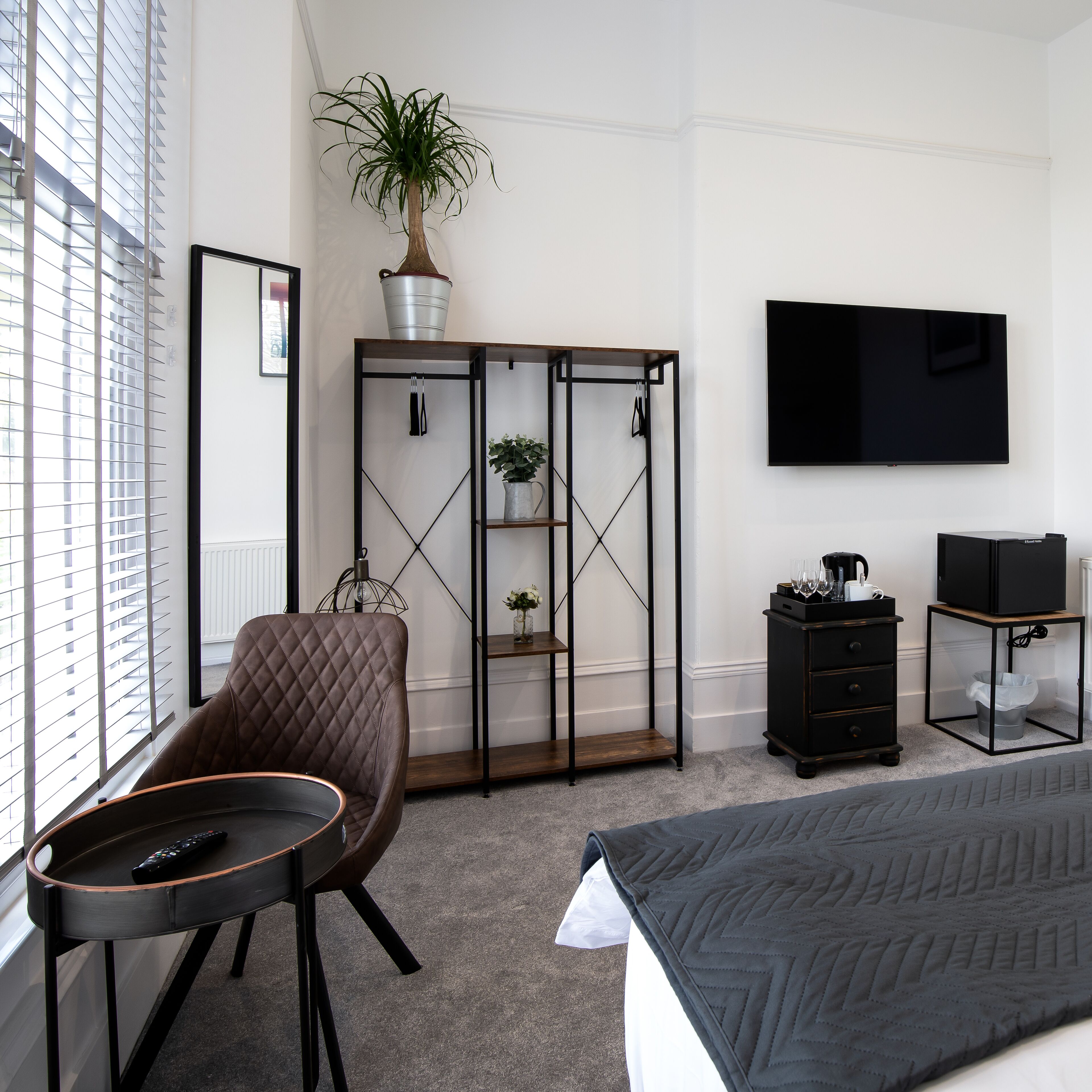 A hotel room with a bed, a chair, a TV, a desk, a plant, and a wardrobe.