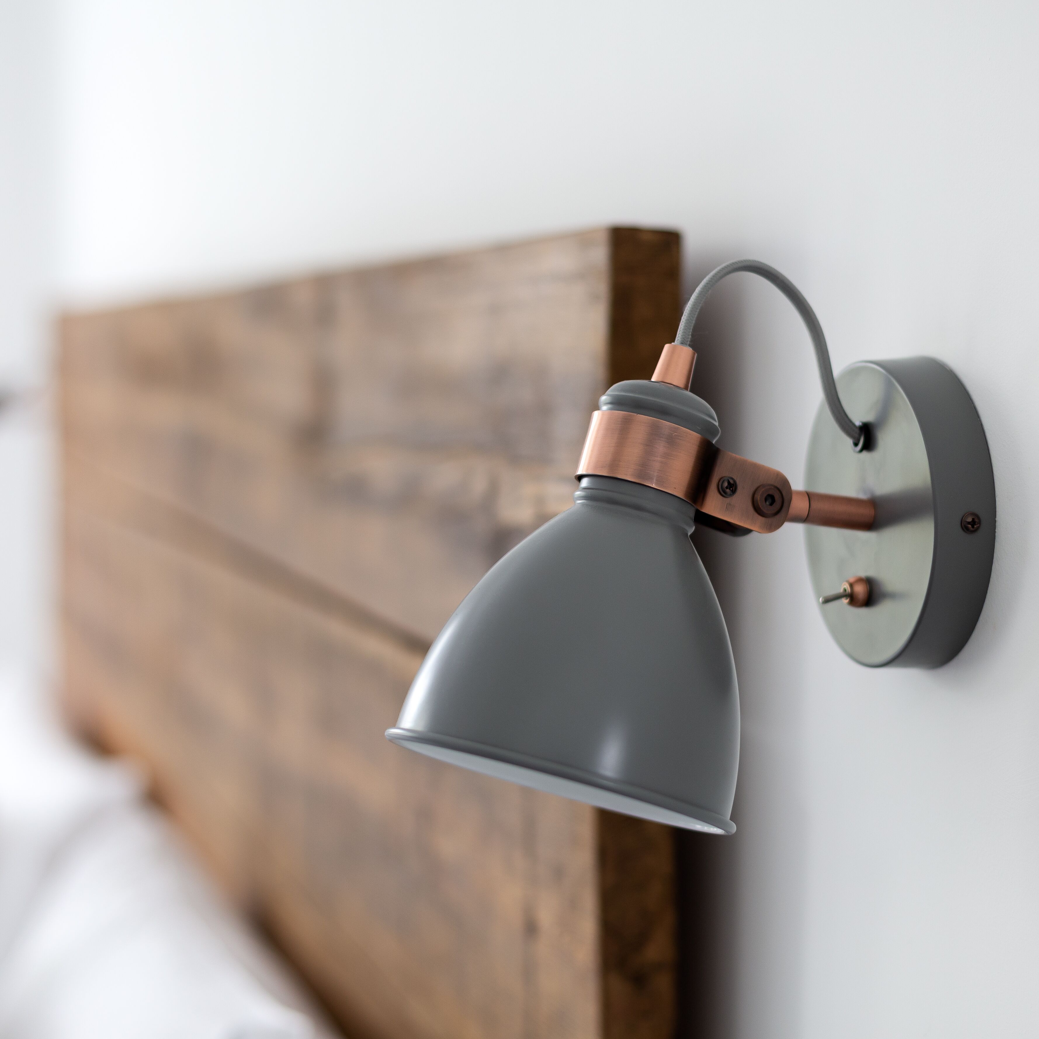 Una lámpara de pared con base de cobre y pantalla gris, fijada a un cabecero de madera.