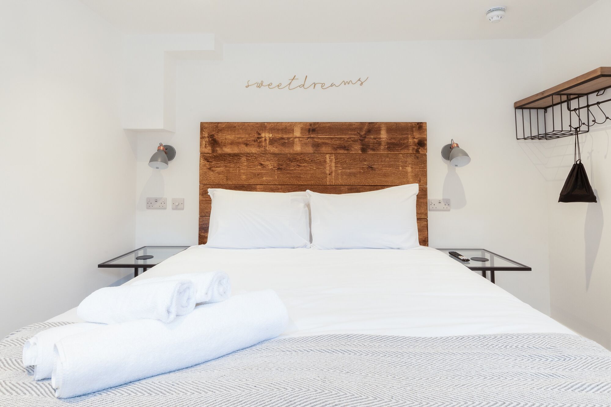 Hotellihuone, jossa on puinen sängynpääty, valkoiset vuodevaatteet, kaksi yöpöytää ja seinään kiinnitetty valaisin.