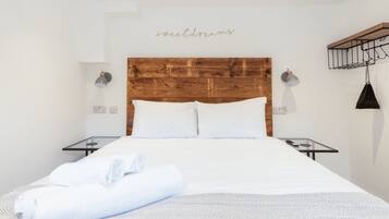 Hotellihuone, jossa on puinen sängynpääty, valkoiset vuodevaatteet, kaksi yöpöytää ja seinään kiinnitetty valaisin.