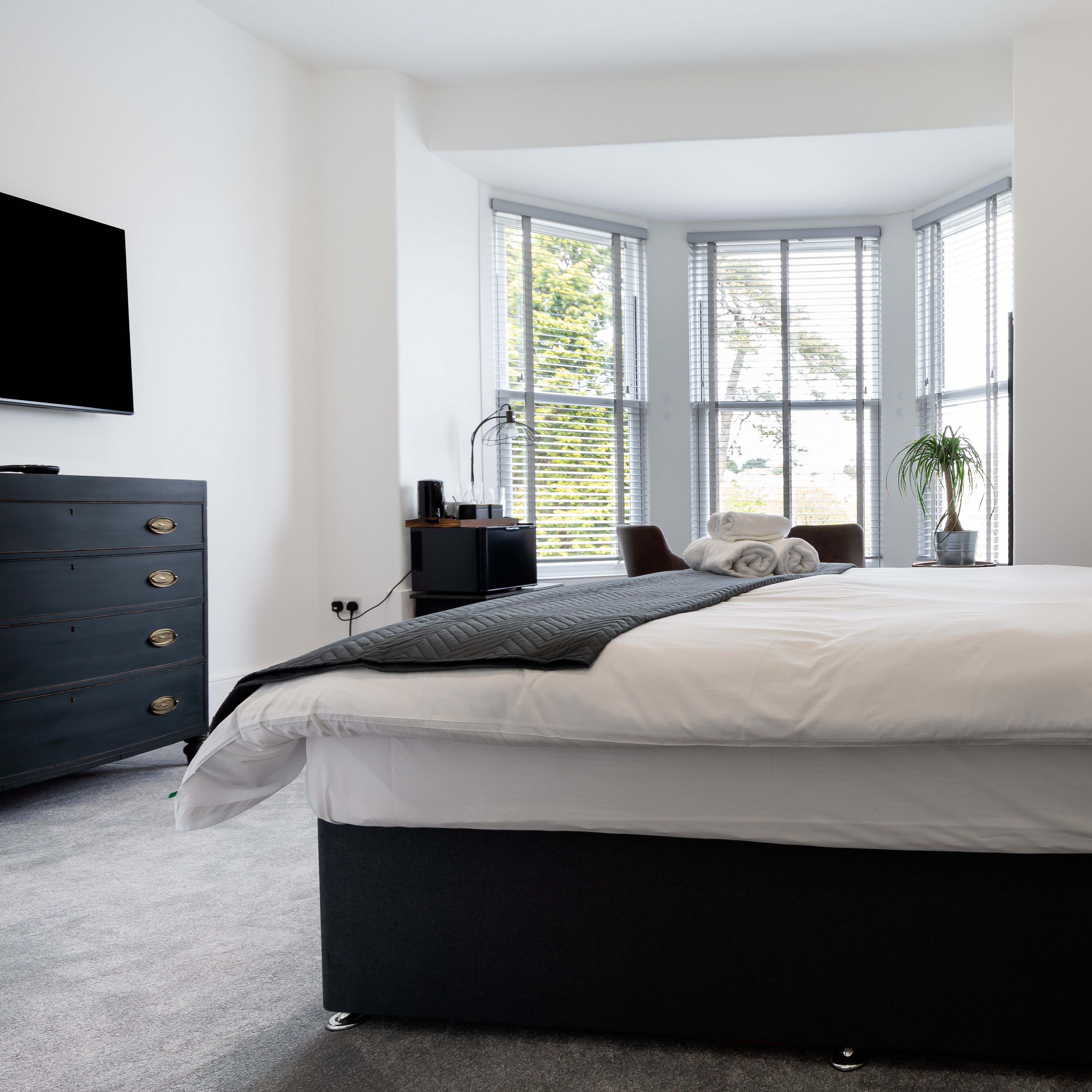 Quarto moderno com cama grande, cômoda escura, TV na parede e janela com persianas.