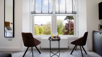 Una habitación moderna con dos sillas, una mesita baja con botellas, un espejo grande y una ventana con persianas.
