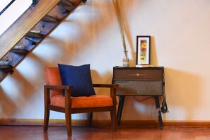 Una habitación con una silla de madera, una mesita con un jarrón, un cuadro enmarco y una escalera.