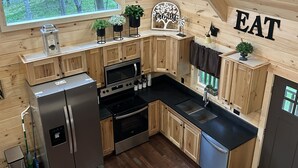 Eine moderne Küche mit Holzschränken, einem Edelstahlkühlschrank, einem Backofen, einer Mikrowelle und einem Spülbecken. Ein Fenster bietet Blick auf Bäume, und ein Schild zeigt das Wort 'EAT'.