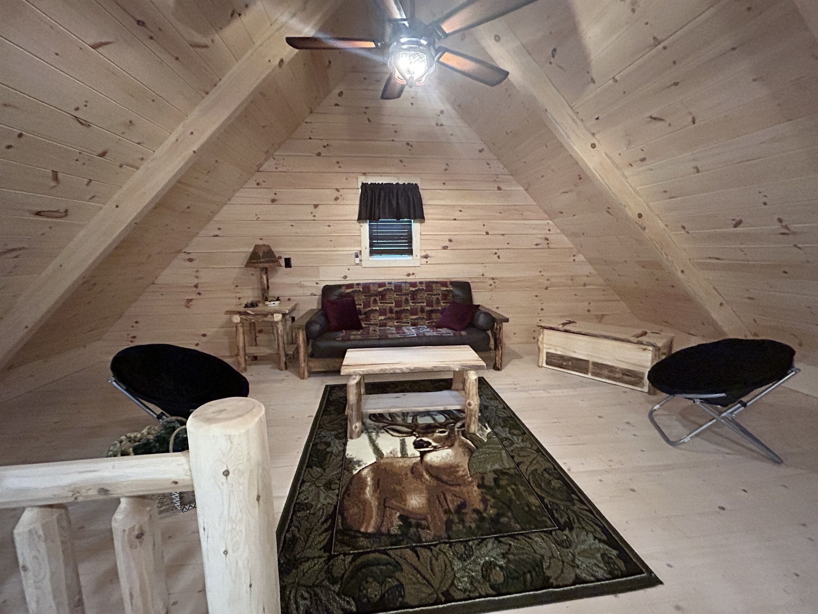 A cozy attic room with a wooden sloped ceiling, a ceiling fan, a sofa, a coffee table, and two chairs.