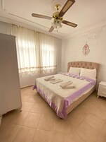 Ein ordentlich bezogenes Bett mit einer violett-weißen Tagesdecke, einem Deckenventilator und einem Fenster mit durchscheinenden Vorhängen.