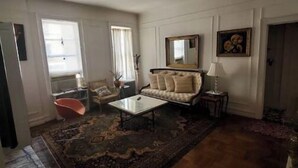 Un salón espacioso con un sofá grande, una mesa de centro y una alfombra estampada. Hay dos ventanas con cortinas, un espejo en la pared y una puerta que conduce a otra habitación.