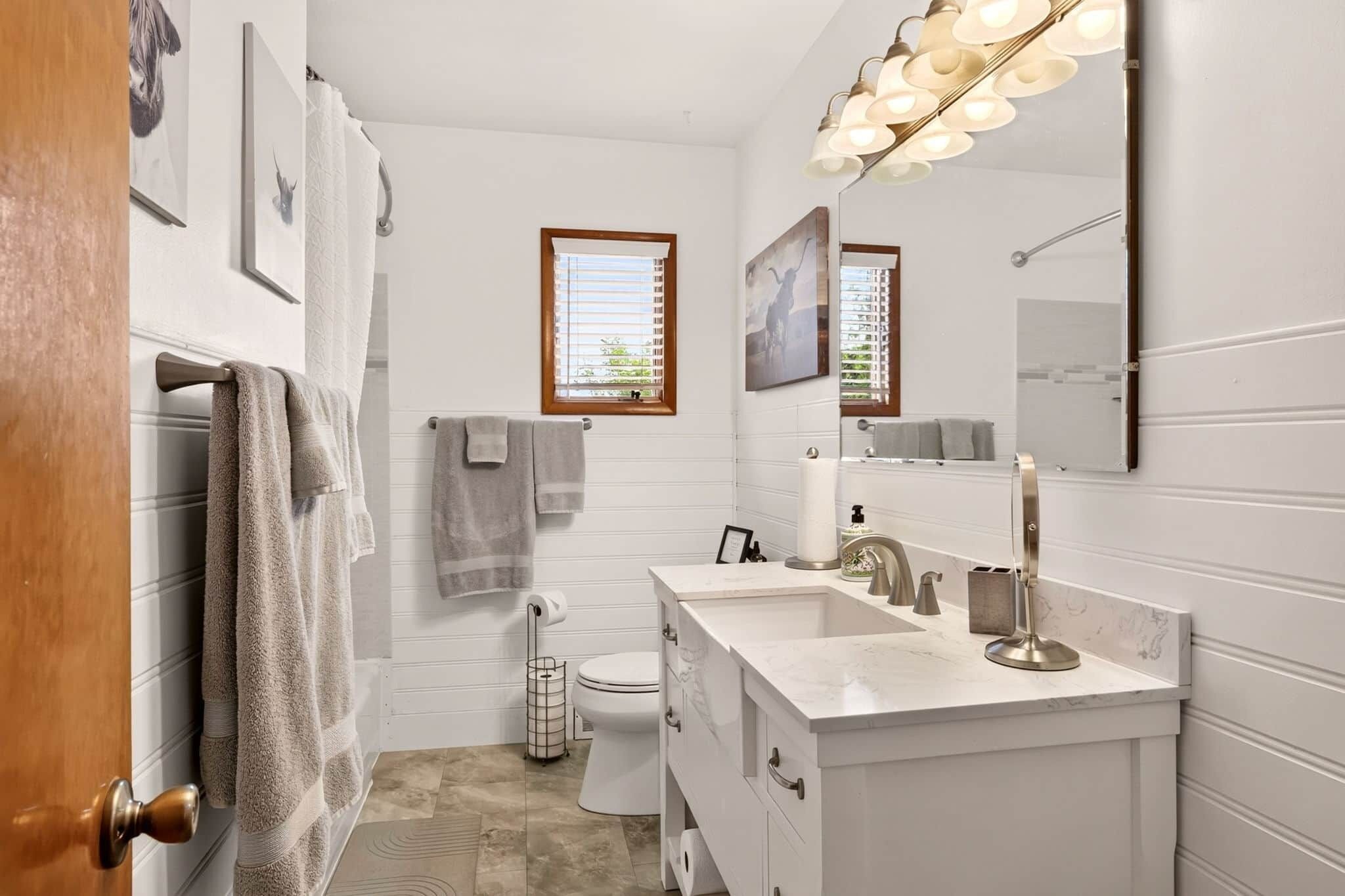 A bathroom with a white vanity, a toilet, a shower area, and a window with blinds.