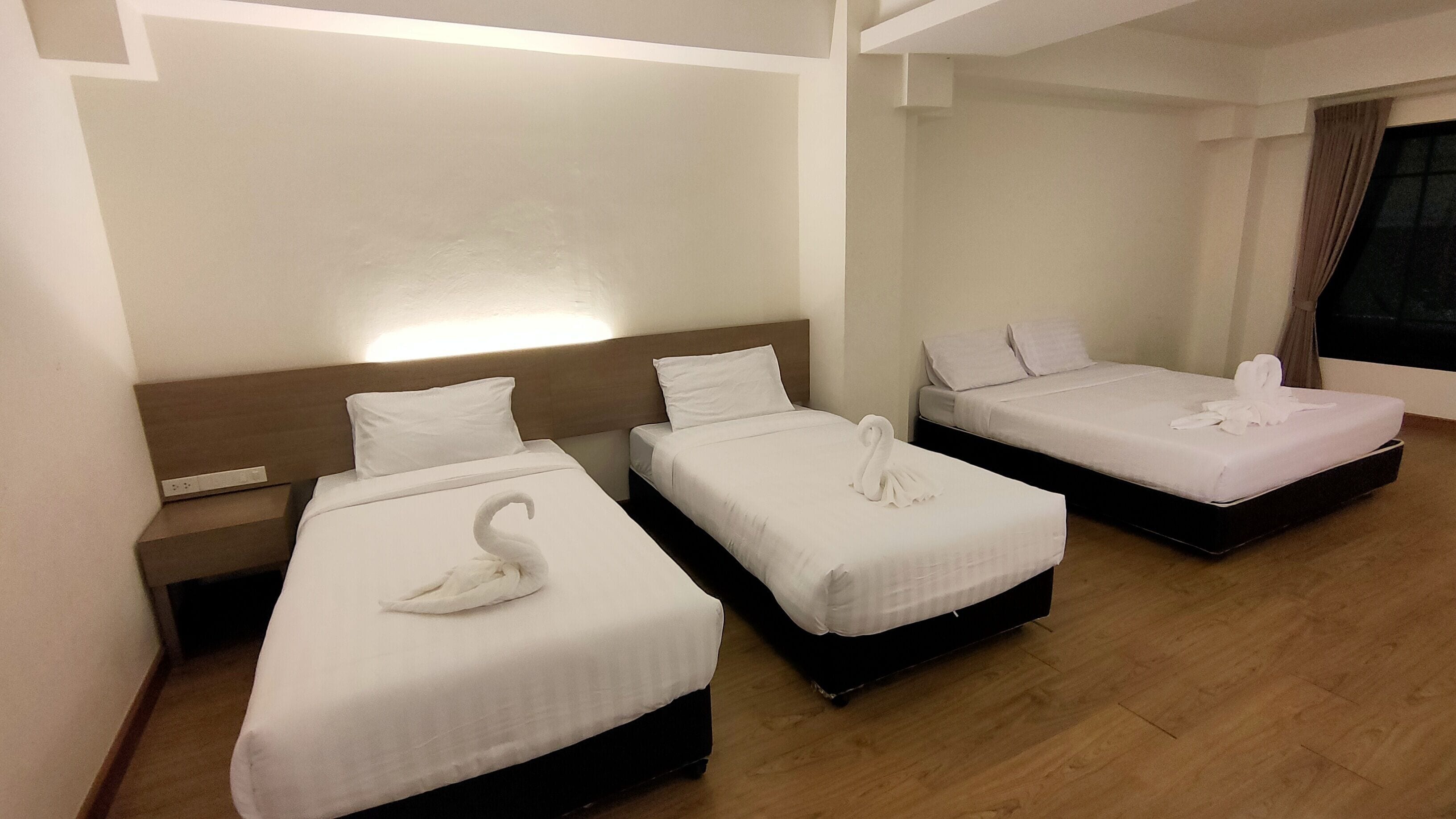 A hotel room with three single beds, each with a white bedspread and pillows. There is a wooden headboard on the left bed, and the room has a wooden floor.