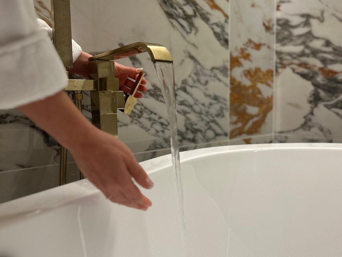 Eine Hand dreht einen Messing-Wasserhahn, während Wasser in eine weiße Badewanne fließt.