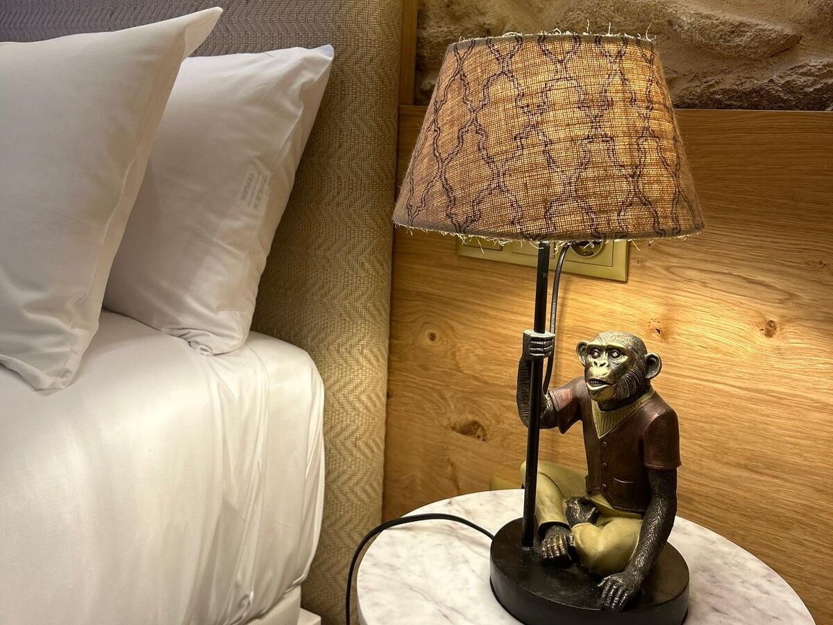 Un lit avec des draps blancs, une lampe dotée d’un abat-jour texturé et une figurine de singe posée sur un socle en marbre.