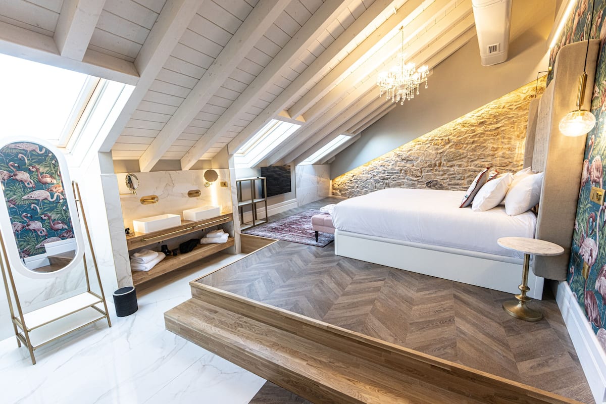Un dormitorio con una cama grande, un muro decorativo de piedra, suelo de madera, tragaluces y un baño con espejo y lavamanos.
