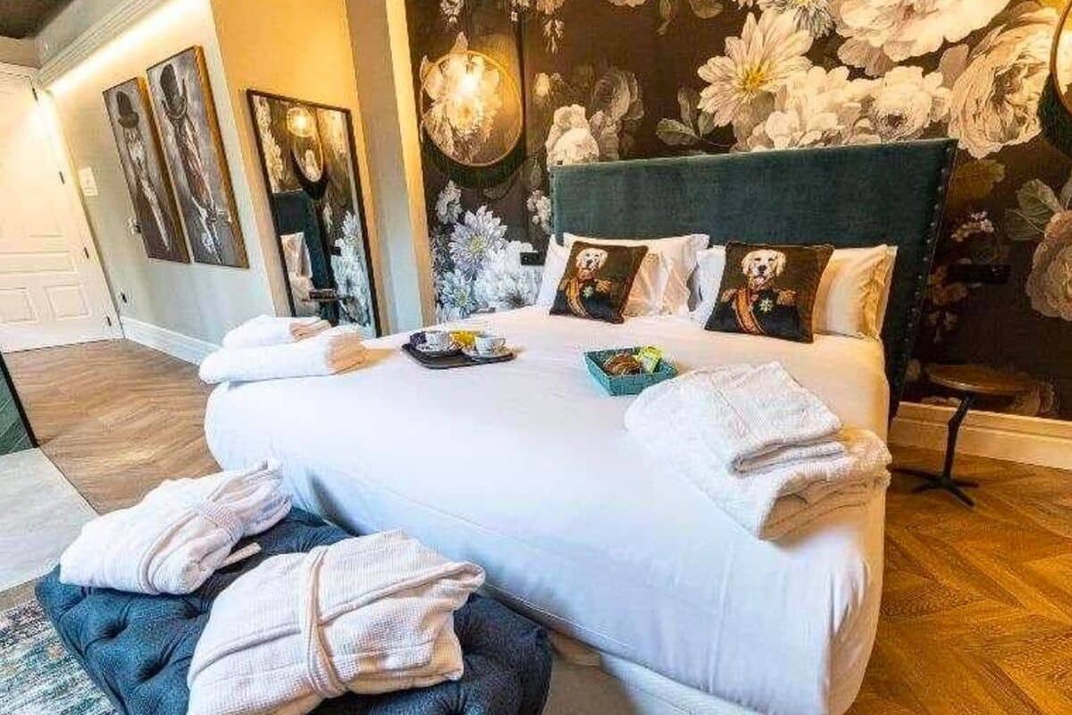 Una cama bien tendida con almohadas y una manta, una mesita de noche con un frutero, y una pared con papel tapiz floral.