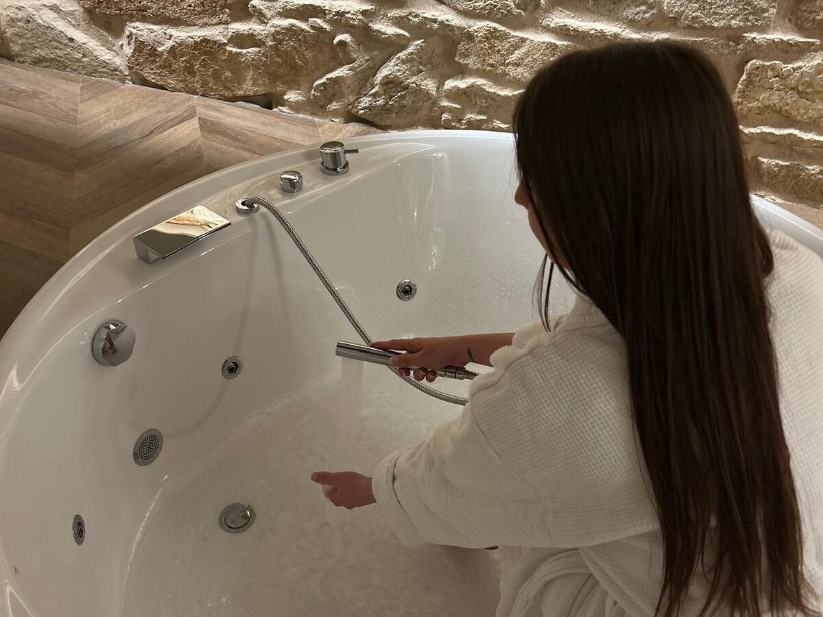 A person in a white robe adjusting a shower head in a large, round bathtub with multiple faucets and a built-in soap dish.
